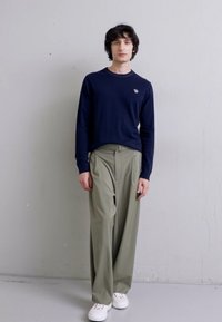 Un pull à manches longues bleu marine, un pantalon large vert olive et des baskets blanches. Le pull présente un petit logo sur la poitrine gauche.