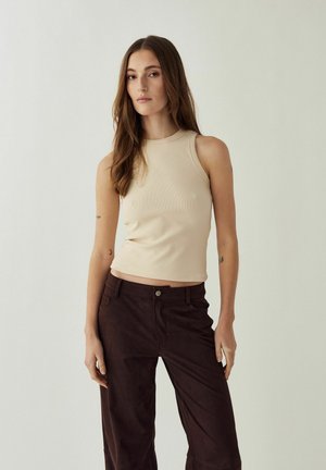 Žena s dlhými hnedými vlasmi, oblečená v bezrukávovom béžovom topu a tmavohnedých nohaviciach, stojaca pred svetlým neutrálnym pozadím.