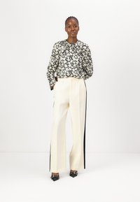 Blumenbluse in Schwarz und Creme, mit langen Ärmeln und runder Ausschnitt. Cremefarbene weite Hosen mit schwarzen Seitenstreifen. Schwarze spitze Schuhe.