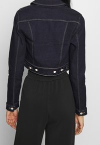 Veste en denim foncé raccourcie avec un col, des accents en bouton et des coutures contrastantes. Elle dispose de manches longues et d'une coupe ajustée.