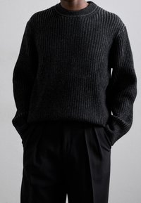 Pull noir en maille côtelée avec un col rond, présentant une apparence texturée. Porté avec un pantalon noir sur mesure.