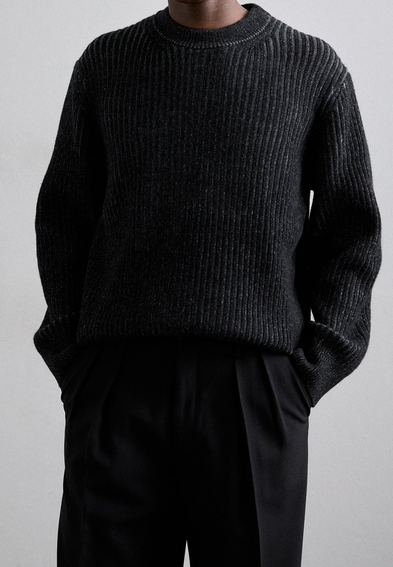 Pull noir en maille côtelée avec un col rond, présentant une apparence texturée. Porté avec un pantalon noir sur mesure.