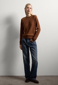 Frau mit langen blonden Haaren steht gegen eine weiße Wand, trägt einen braunen Pullover, blue Jeans und dunkle Schuhe, mit einer Hand in der Tasche.