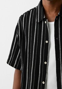 Camicia nera a maniche corte con strisce verticali bianche, tessuto testurizzato e chiusura a bottoni con bottoni di colore chiaro.