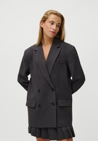Blazer cinza de corte duplo com lapelas com corte, bolsos e botões pretos, combinado com uma saia plissada, apresentando uma textura suave.