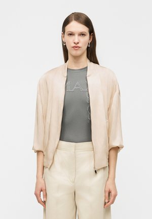 Pikkade pruunide juustega naine kannab beežikat bomberjopet, halli soonikkoes topi nõrga tekstiga ja kõrge vöökohaga kreemivärvi pükse, taustaks on lihtne taust.