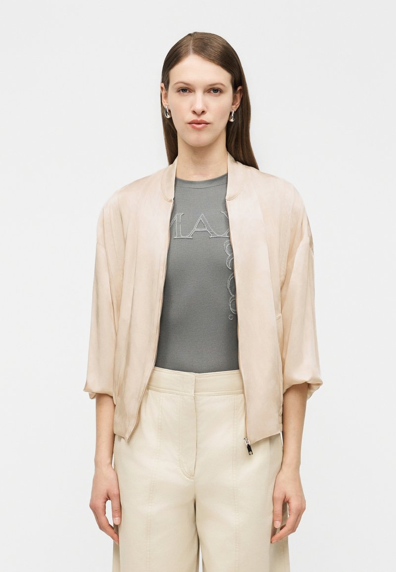 Γυναίκα με μακριά καστανά μαλλιά φοράει μπεζ μπουφάν bomber, γκρι ριμπ τοπ με διακριτικό κείμενο και ψηλόμεσο κρεμ παντελόνι, στέκεται μπροστά σε απλό φόντο.