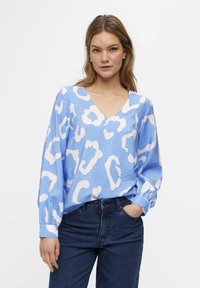 Kvinde med langt hår iført en blå og hvid mønstret V-hals bluse og mørkeblå jeans, den ene hånd i lommen, neutral baggrund.