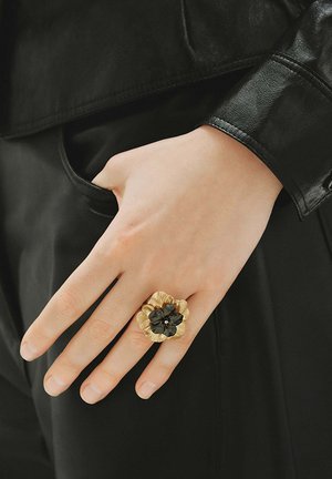 Mano con anillo dorado y negro en forma de flor descansando cerca de un bolsillo, con chaqueta de cuero negra y pantalones negros.