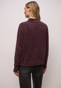 Lila Strickpullover mit hohem Kragen und geripptem Saum. Verfügt über eine strukturierte Oberfläche und angesetzte Schultern, kombiniert mit grauen Hosen.