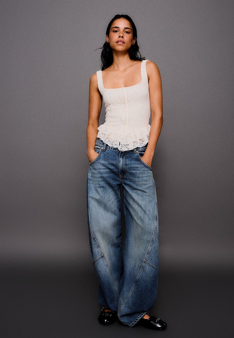 Femme aux cheveux foncés porte un haut blanc en dentelle sans manches, un jean large bleu et des chaussures noires, debout les mains dans les poches sur fond gris.