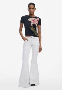 Schwarzes T-Shirt mit floralem Print auf der linken Seite, kombiniert mit hoch taillierten, ausgestellten weißen Jeans. Das Modell steht mit einer Hand in der Tasche.