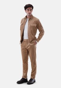 Beige Kordhemdjacke mit frontalen Taschen, kombiniert mit passenden Hosen. Darunter ein weißes T-Shirt und schwarze Schnürschuhe. Glatte Textur.