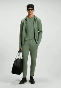 Athletic outfit in muted green fabric, featuring a fitted top, zip-up jacket, and tapered pants with slits. Accompanied by a black duffel bag.