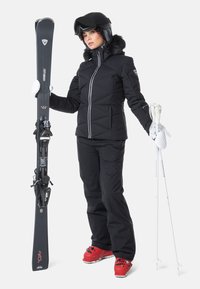 Svart skidjacka med vita detaljer, svarta skidbyxor, röda skidor, håller svarta skidor och vita stavar. Ytan är slät och vadderad.