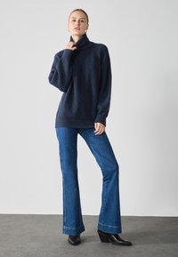 Pulover tricotat de culoare bleumarin cu guler înalt, asortiat cu blugi evazați de culoare bleumarin închis și ghete cu toc negre pe un pardoseală gri.