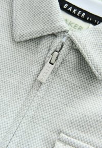 Harmaa kuvioitu kauluspaita, jossa on vetoketju ja korostettu vetotappi. Kangas on tekstuuriltaan ruudukkomainen.