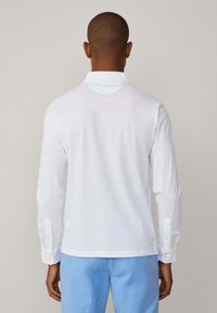Langärmliges weißes Poloshirt mit umgeschlagenem Kragen, geknöpften Manschetten und einem schlichten Rückendesign, kombiniert mit hellblauen Hosen.