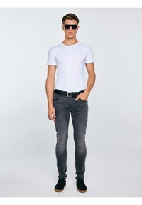 Fiatal férfi áll, fekete napszemüveget visel, fehér rövid ujjú henley ing, fakó szürke farmernadrág, fekete öv és fekete, gumitalpú sportcipő van rajta.