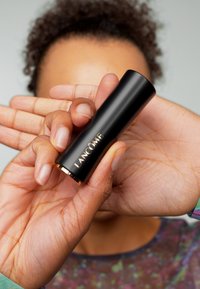 Svart cylindrisk läppstiftförpackning med gulddetaljer och "Lancôme"-logotyp, hålls i hand. Bakgrunden består av mjuka hudtoner och suddiga färger.