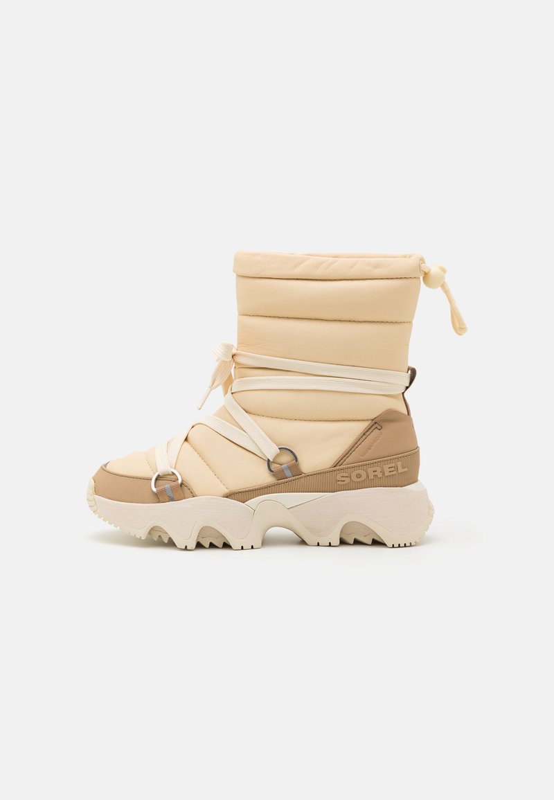 Botte rembourrée beige avec un bout rond, des sangles élastiques et des anneaux en métal. Matériau texturé, semelle extérieure blanche chunky avec motif en zigzag.