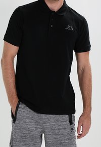 Svart polotröja med krage, två knappar och en liten broderad logotyp på vänster bröst, ihop med grå shorts.