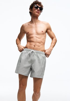 Mężczyzna bez koszulki w czarnych okularach przeciwsłonecznych i szarych kąpielówkach w kratę z sznurkiem, pozujący na białym tle.