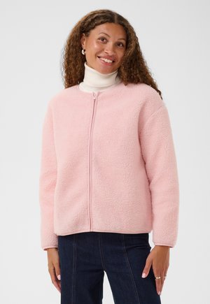 Femme souriante, portant une veste polaire rose clair zippée sur un col roulé blanc et un jean bleu foncé, debout devant un fond uni.
