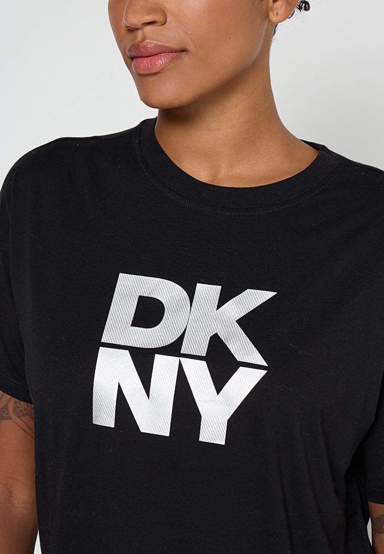 Femme portant un T-shirt noir avec un grand logo DKNY blanc sur la poitrine, montrant un gros plan partiel des lèvres au torse.