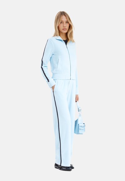 Tuta azzurro chiaro con strisce nere, dotato di giacca con zip e pantaloni con vita elastica. Tessuto morbido e vestibilità comoda. Piccola borsa laterale.