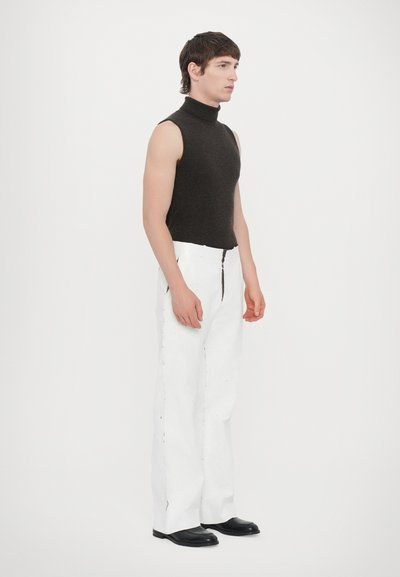 Fitted sleeveless turtleneck in dark brown wool, paired with white wide-leg trousers featuring black zipper accents. Black footwear completes the look.