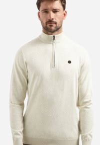 Licht beige pullover met een halve rits aan de voorkant, geribbelde manchetten en onderrand. Beschikt over een klein hexagonaal logo op de borst. Zachte gebreide textuur.