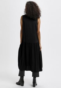 Robe noire sans manches avec une jupe à volants, présentant une texture douce et une coupe décontractée, associée à des bottines noires et un pantalon noir.