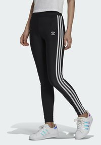 Leggings negri cu trei dungi albe pe laterale, având un mic logo Adidas pe partea superioară a piciorului și un design ajustat.