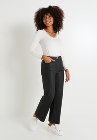 Mujer con cabello rizado sonriendo, vestida con una blusa blanca de manga larga con encaje, pantalones anchos negros y zapatillas blancas, de pie contra una pared blanca.