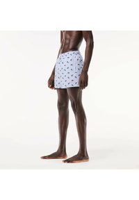 Costumi da bagno da uomo bianchi con una trama di foglie verdi ripetuta, vita elastica, tasche laterali e vestibilità rilassata.