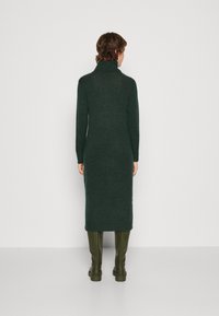 Garšs, tumši zaļš adīts kleita ar augstu turtleneck apkakli un garām piedurknēm. Kombinēta ar ceļgala augstuma zaļām zābakām. Strukturēta auduma, pieguļošs siluets.