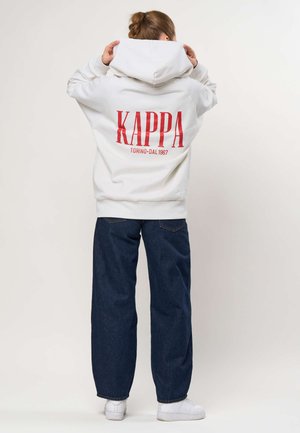 Asmuo dėvintis baltą džemperį su raudonu užrašu „KAPPA TORINO DAL 1967“ nugaroje, mėlynais džinsais ir baltais sportbačiais, reguliuojantis gaubtą.