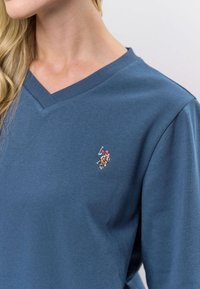 Tumši zils kokvilnas V-veida kakla krekls ar nelielu izšūtu logotipu uz kreisā krūtīm. Gluda tekstūra ar ribotu apkakles apdari.