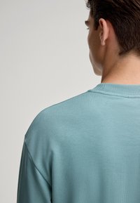 Felpa blu chiaro con colletto a coste e cuciture sulle spalle, caratterizzata da una superficie di tessuto liscia e una vestibilità rilassata. Vista posteriore visibile.