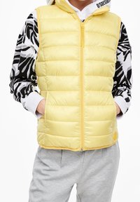 Žlutá péřová vesta s prošívanou strukturou, s plným zipem a bočními kapsami, oblečená přes mikinu s černo-bílým vzorem. Šedé tepláky.