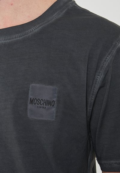 T-shirt in cotone grigio scuro con scollatura rotonda, caratterizzato da un patch rettangolare con "Moschino Swim" in lettere nere. Texture liscia.