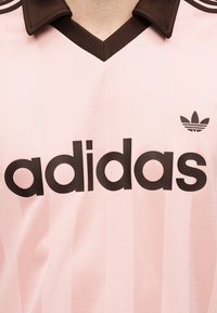 Ανοιχτό ροζ μπλουζάκι Adidas με μαύρο γιακά, μαύρο λογότυπο Adidas και έντονο μαύρο κείμενο "adidas" στο μπροστινό μέρος.