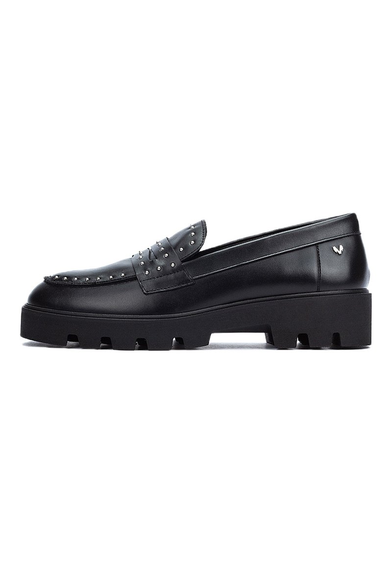 Mocasines de cuero negro con acentos de tachuelas, una suela de plataforma gruesa y una textura suave. Presenta un detalle de lazo decorativo en la parte frontal.