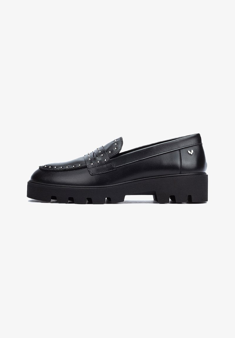 Mocasines de cuero negro con acentos de tachuelas, una suela de plataforma gruesa y una textura suave. Presenta un detalle de lazo decorativo en la parte frontal.