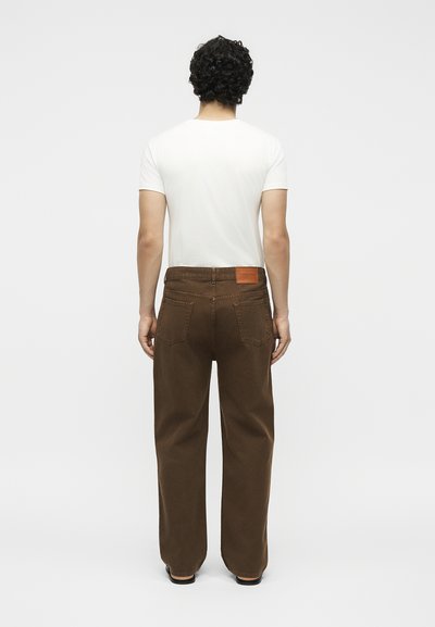 Άνδρας με σγουρά σκούρα μαλλιά που φοράει απλό λευκό κοντομάνικο πουκάμισο και φαρδιά καφέ παντελόνια, στέκεται πλάτη σε απλό φόντο.