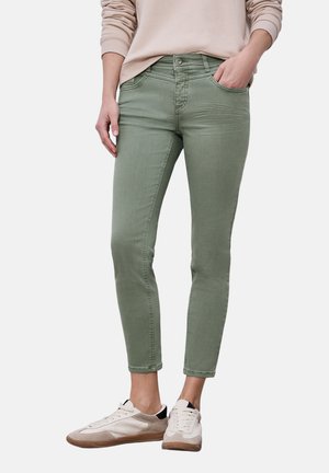 Person trägt hellgrüne Slim-Fit-Jeans, beige Sweatshirt und weiße Turnschuhe, steht mit einer Hand in der Tasche vor weißem Hintergrund.