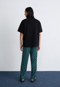 En person med langt hår bærer en sort oversized t-shirt og mønstrede grønne bukser, stående opad en lys væg og et mørkt tæppe.