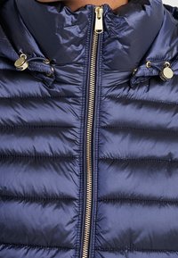 Veste matelassée bleu marine avec fermeture éclair dorée et cordons de serrage réglables sur le col montant, portée par une personne.