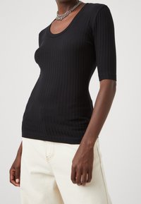 Černé proužkované tričko s krátkými rukávy a kulatým výstřihem, vyrobené z elastického materiálu, v kombinaci se světlými kalhotami.